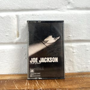Puede incluir: Un casete vintage de Joe Jackson "Look Sharp!". La caja del casete es negra con texto blanco y un gráfico de silueta. El logotipo de A&M Records es visible. El casete está sobre una superficie de madera.