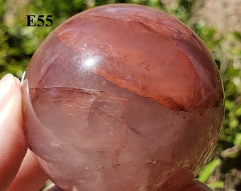 May include: A clear, spherical crystal ball with reddish-brown inclusions and a polished surface. The crystal is held in a hand, with the text "E55" visible on the surface. The crystal ball is displayed against a blurred green background.