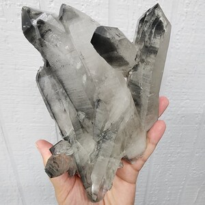 May include: A cluster of clear quartz crystals with black inclusions. The crystals are arranged in a point-up formation, with the largest crystal at the top. The crystals are translucent and have a glassy luster.