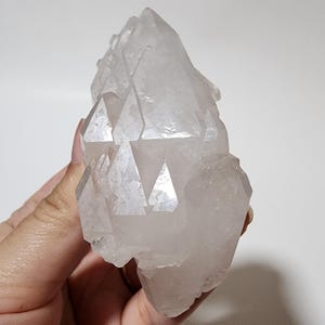 May include: A clear quartz crystal cluster with multiple points and facets. The crystal is held in a hand, showcasing its natural, irregular shape and translucent appearance. The quartz appears to be unpolished, highlighting its raw, natural beauty.