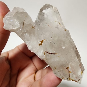 Puede incluir: Un grupo de cristales de cuarzo transparente con inclusiones de rutilo dorado. El cristal tiene una textura natural rugosa. El cuarzo es transparente, permitiendo el paso de la luz, y las agujas de rutilo añaden interés visual. El cristal se sostiene en una mano.