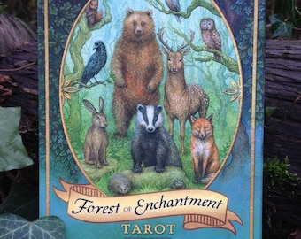 May include: A tarot card deck titled "Forest of Enchantment" featuring illustrations of forest animals like a bear, deer, fox, and owl. The deck has a teal border with gold accents and the author's and illustrator's names.