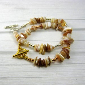 May include: A multi-strand bracelet featuring irregular, polished shell chips in shades of cream, tan, and brown. The design incorporates small metallic gold-tone spacer beads and is secured with a gold-tone toggle clasp closure at the front.