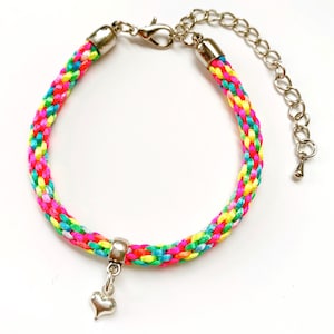 May include: A colorful braided bracelet with a silver heart charm and a silver lobster clasp. The bracelet is made of multiple strands of brightly colored thread, including pink, yellow, green, and blue.