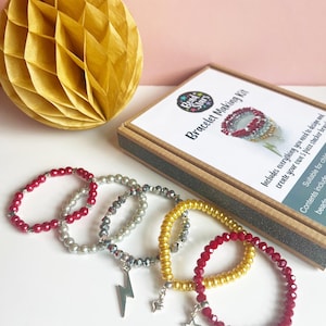 May include: A bracelet-making kit with several beaded bracelets in red, silver, and gold. Some bracelets have charms, including a lightning bolt and a star. The kit box is visible, with the text "Bracelet Making Kit". A yellow honeycomb paper decoration is in the background.