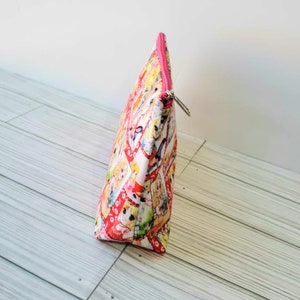 May include: A pink zippered pouch with a colorful cartoon print. The pouch is made of fabric and has a white background with a variety of cartoon characters in bright colors.