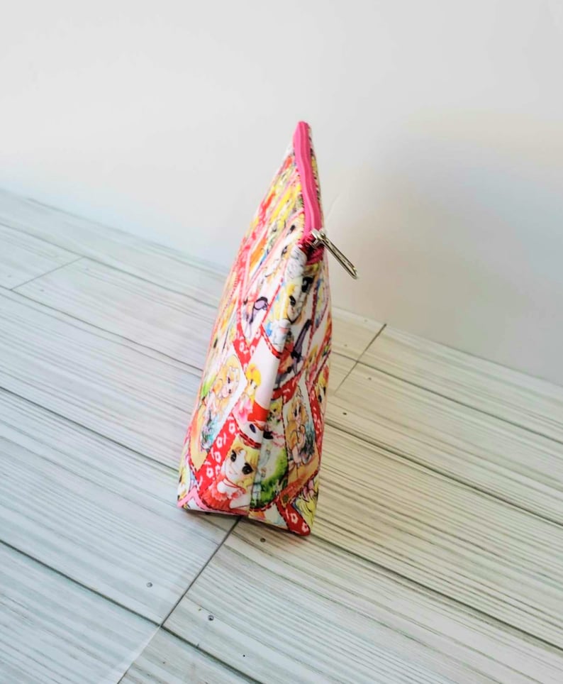 May include: A pink zippered pouch with a colorful cartoon print. The pouch is made of fabric and has a white background with a variety of cartoon characters in bright colors.