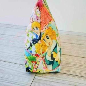 May include: A pink and white zippered pouch with a colorful cartoon print featuring two girls with blonde hair. The print includes flowers, a house, and other details.