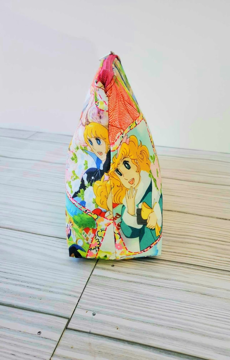 May include: A pink and white zippered pouch with a colorful cartoon print featuring two girls with blonde hair. The print includes flowers, a house, and other details.