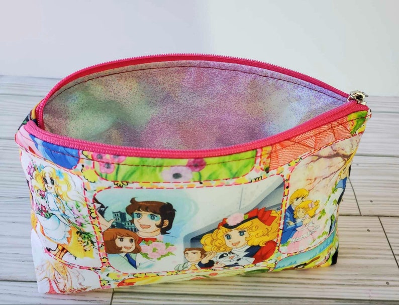 May include: A pink zippered pouch with a colorful floral print featuring cartoon characters. The pouch is open and the inside lining is pink.