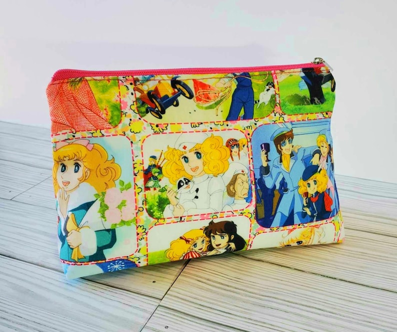 May include: A pink zippered pouch with a colorful patchwork design featuring cartoon characters and images. The pouch is made of fabric and has a white background with pink, blue, yellow, and green accents.
