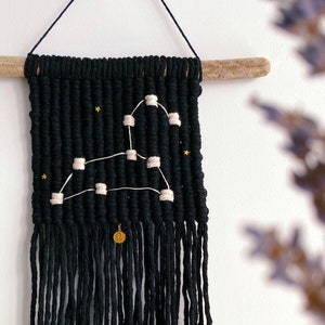 Peut inclure: Suspension murale en macramé noir représentant la constellation du Lion en fil crème. L'œuvre est suspendue à un morceau de bois flotté et comprend des étoiles dorées et un petit charme doré. Affiché sur un mur blanc.