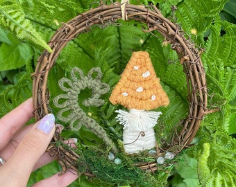 以下が含まれることがあります： 小枝でできた小さなリースに、キノコとシダが刺繍されています。リースは苔と小さな白いビーズで飾られています。
