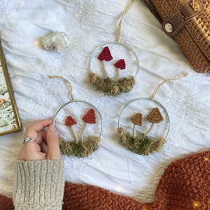 Puede incluir: Tres adornos de crochet en forma de hongo que cuelgan de aros plateados. Cada adorno presenta dos hongos con sombreros de diferentes colores y un lecho de musgo verde.
