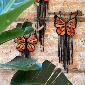 May include: Three macrame wall hangings featuring orange and brown butterflies with black fringe. The butterflies are made of yarn and are hanging on a brick wall.