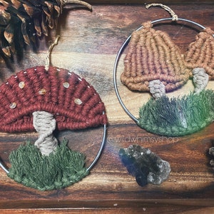 Puede incluir: Dos tapices de pared de macramé con forma de hongo. Uno es rojo con puntos blancos y el otro es marrón. Ambos tienen hierba de macramé verde en la parte inferior.