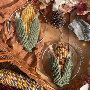 Puede incluir: Dos decoraciones de pared de maíz hechas a mano con cáscaras verdes y granos amarillos. El maíz está envuelto en un aro de alambre plateado y colgado en una superficie de madera.