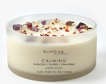 Puede incluir: Una vela de cera de soja blanca con cristales de amatista, pétalos de rosa y cristales de cuarzo transparentes. La vela está en un tarro de vidrio transparente y tiene el texto "Gum Drop Australia" y "CALMING Protection | Humility | Stress Relief AMETHYST Crystal Infused Soy Wax Candles" en la etiqueta.