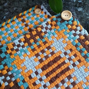 May include: Two handmade woven potholders in a colourful pattern. The design features a grid of brown, orange, and teal blue squares. Each potholder has a wooden button that reads "handmade".