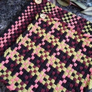 Op de afbeelding: Twee geweven pannenlappen in een dambordpatroon. De kleuren zijn zwart, roze, groen en bordeauxrood. Elke pannenlap heeft een houten knoop. De pannenlappen zijn handgemaakt en geschikt voor gebruik in de keuken.