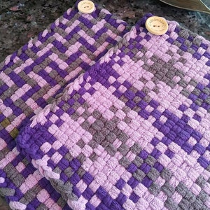 Può includere: Due presine fatte a mano, tessute in tonalità di viola, lavanda e grigio. Le presine presentano un motivo a trama a cestino e sono fissate con un bottone di legno con la scritta "handmade".