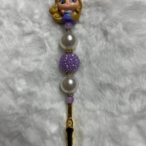 May include: A decorative hair clip featuring a cartoon princess figure with blonde hair and a blue dress. The clip has a gold-colored metal base, adorned with white pearl beads and a sparkling purple bead.