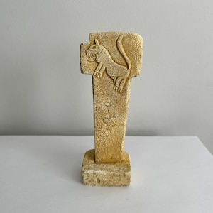 May include: A beige stone sculpture of a stylised animal with a long tail. The sculpture is on a square base and has a textured surface.