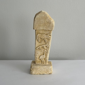 May include: A small, beige stone sculpture of a standing figure with carvings of animals. The sculpture has a rounded top and a rectangular base.