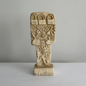 Puede incluir: Una pequeña escultura de piedra blanca tallada que representa una figura con tres arcos en la parte superior y diseños intrincados en la parte delantera. La escultura está sobre una pequeña base cuadrada.