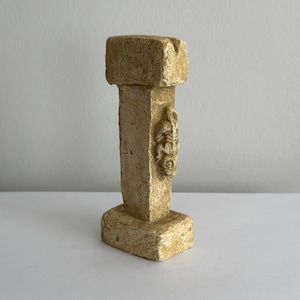 May include: A small, beige, stone sculpture of a column with a decorative design on the side. The column has a square base and a square top.
