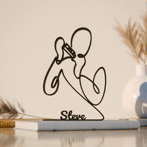 Puede incluir: Una taza de cerámica blanca con asa negra y el texto "But First, Coffee" en negrita negra. La taza se encuentra sobre un fondo borroso, sugiriendo un ambiente acogedor.