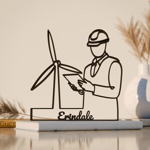 Könnte beinhalten: Schwarze Metallskulptur, die einen Ingenieur mit Windrad und Tablet darstellt, mit dem Namen "Erindale". Das Kunstwerk steht auf Büchern, mit einer Vase und einer dekorativen Pflanze im Hintergrund. Dekorativer Gegenstand.