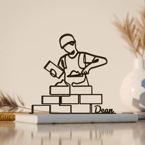 May include: Black metal sculpture depicting a bricklayer building a wall. The artwork shows a minimalist line drawing of a person wearing a hard hat and holding a trowel. The name "Dean" is written on the base.