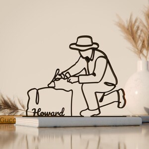 Pode incluir: Escultura em metal preto que retrata uma pessoa ajoelhada escrevendo em uma superfície. A escultura é um design de linha contínua, com o nome "Howard" escrito na superfície. A escultura é colocada em um livro.