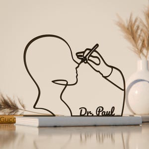 Op de afbeelding: Zwarte metalen sculptuur die een gezicht afbeeldt dat wordt bewerkt door een hand die een gereedschap vasthoudt. De sculptuur is gemonteerd op een grijs boek, met de naam "Dr. Paul" in cursief geschreven. Een vaas met gedroogde planten staat op de achtergrond.