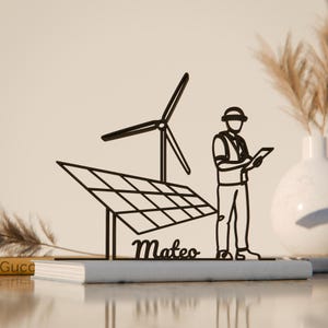 Könnte beinhalten: Schwarze Metallskulptur, die ein Solarpanel, eine Windturbine und einen Ingenieur mit einem Tablet darstellt. Der Name "Mateo" ist kursiv geschrieben. Die Skulptur steht auf einem grauen Buch.