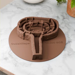 Peut inclure: Modèle marron de Göbeklitepe, site historique, avec les inscriptions "Göbeklitepe" et "10.000 BCE" en relief sur la base. Le modèle présente des éléments architecturaux détaillés, posé sur une surface en marbre.