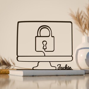 Op de afbeelding: Zwart metalen silhouet van een computermonitor met een hangslotpictogram. De monitor staat op een grijs boek, met de naam "Jackie" in cursief. Een witte vaas en gedroogde planten staan op de achtergrond.