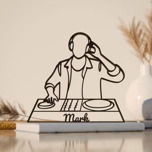 Könnte beinhalten: Schwarze Metall-DJ-Silhouette mit Kopfhörern, Händen an einem Mischpult und dem Namen "Mark" darunter. Das Kunstwerk befindet sich auf einer hellen Oberfläche, mit einem Buch und einer dekorativen Vase im Hintergrund.