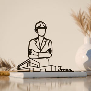 Pode incluir: Desenho a traço preto de um arquiteto com os braços cruzados, usando um capacete, em frente a um projeto de edifício. O nome "Jenna" está escrito em itálico. A arte é colocada em um livro cinza.