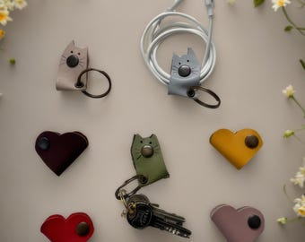 May include: A collection of leather cord organisers and key holders in various shapes and colours. Some are cat-shaped with snap buttons and metal rings, while others are heart-shaped. One cat organiser holds a white charging cable.