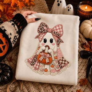 May include: A white sweatshirt featuring an embroidered ghost character wearing a large pink leopard and gingham print bow. The ghost holds an orange pumpkin basket decorated with a small bow and is surrounded by small pumpkins and floral patterns.