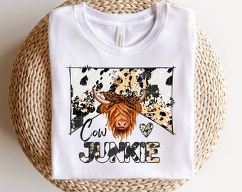 Puede incluir: Camiseta blanca con un gráfico de vaca negro, blanco y marrón con un pañuelo. El texto "Cow Junkie" está debajo del gráfico de la vaca.