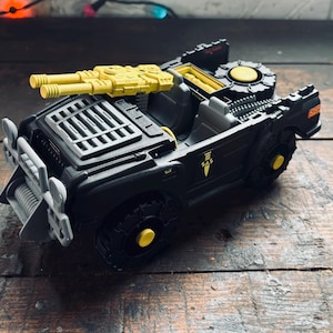 May include: A black toy jeep with yellow accents, including a double-barreled gun on top and wheel details. The jeep features a winch, a grill, and a rear-mounted circular structure. The wheels and other details are yellow. The jeep is on a wooden surface.