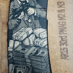 May include: A black AR15 30rd PMAG M2 & M3 magazine featuring a laser-engraved graphic of a man in a pinstripe suit holding a rifle over stacks of hundred-dollar bills. The magazine surface displays detailed line art of currency and the character.