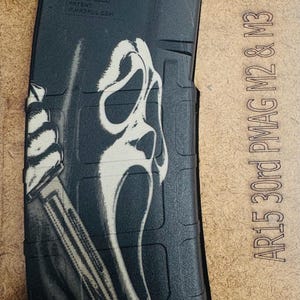 Puede incluir: Un cargador de rifle de polímero negro con un gráfico blanco de una figura enmascarada que sostiene un cuchillo. El cargador tiene texto en relieve que incluye 5.56x45, PMAG 30 y Magpul Industries Corp. El texto AR15 30rd PMAG M2 & M3 está impreso cerca.