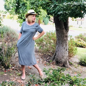 Puede incluir: Una mujer con un vestido midi gris, un sombrero de paja y zapatos planos, de pie frente a un &aacute;rbol.