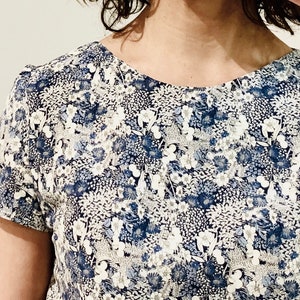 Puede incluir: Una camisa blanca de manga corta con un estampado floral azul. La camisa tiene un cuello redondo y un corte holgado.