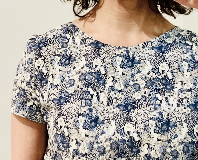 Puede incluir: Una camisa blanca de manga corta con un estampado floral azul. La camisa tiene un cuello redondo y un corte holgado.