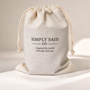 May include: A beige drawstring bag with the text "SIMPLY SAID CO." and the tagline "- Inspired by words that stay with you." The bag is made of a textured fabric and has a natural rope drawstring.